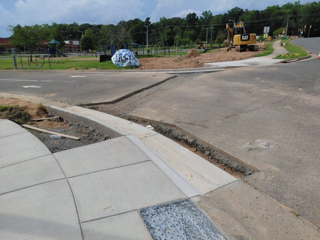 New asphalt patch tying into a fresh concrete curb ramp at a road intersection, with excavators working in the background
