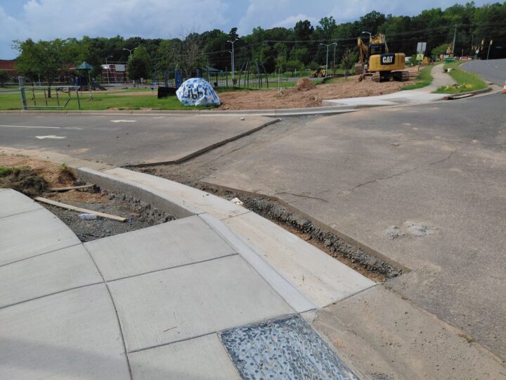 New asphalt patch tying into a fresh concrete curb ramp at a road intersection, with excavators working in the background